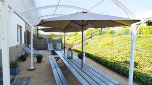 Un patio couvert, doté de bancs en bois et d’un grand parasol, est entouré d’un jardin et d’un escalier menant à un niveau supérieur.