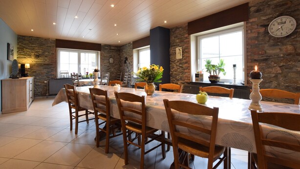 Un coin repas avec une longue table, des chaises, une horloge au mur et une fenêtre ornée de plantes.