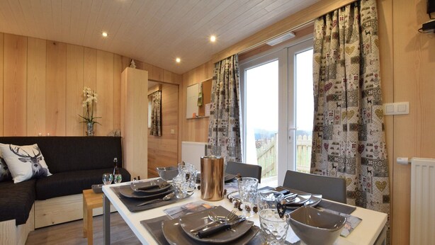 A dining area with a table set for four, wooden walls, a window with patterned curtains, and a view of a fence outside. - Tranquil Stay in Vielsalm (Vielsalm)