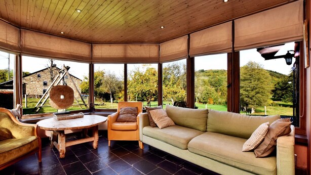 A cozy room with a wooden ceiling, a green sofa, a wooden table, and a chair. Large windows offer a view of a garden and a stone building.