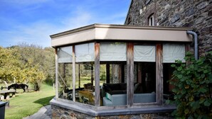 A covered outdoor seating area with stone walls and glass panels, overlooking a grassy yard and a building with a stone facade.