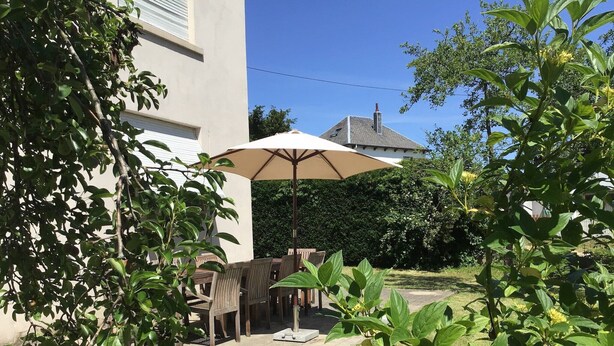 Een terras buiten met een tafel en stoelen onder een grote parasol, omringd door groen en een gebouw.