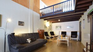 Een gezellige woonkamer met een donkergrijze bank, twee zwarte fauteuils, een houten salontafel en een houten vloer. Daarboven is een balkon met een blauwe balustrade zichtbaar.