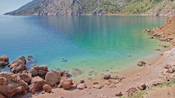 A coastal landscape with a clear lake, rocky shoreline, and a mountain in the background.