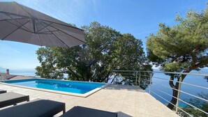 A swimming pool with lounge chairs and an umbrella, surrounded by trees and overlooking a body of water.