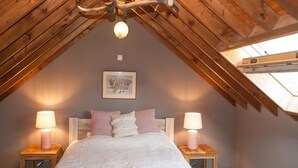 A cozy attic bedroom with a sloped ceiling, a chandelier, a bed with patterned bedding, two bedside tables with lamps, and a framed picture on the wall. - Cozy Forest Cottage in Awenne (Awenne)