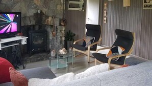 A cozy living room with a stone wall, a fireplace, two armchairs, a glass coffee table, and a television.