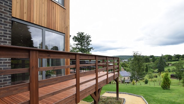 Eine Holzterrasse, an ein modernes Gebäude mit großen Fenstern angebaut, mit Blick auf eine grüne Landschaft.