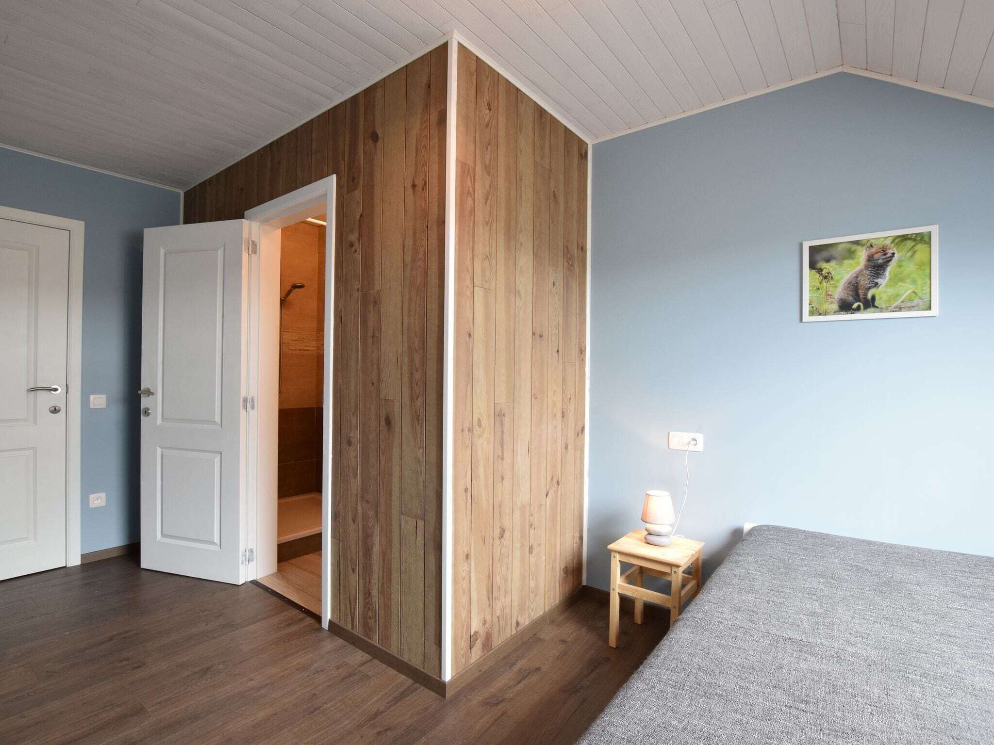 Una habitación con un separador de madera, una puerta que da al baño, una mesita con una lámpara y un cuadro en la pared.
