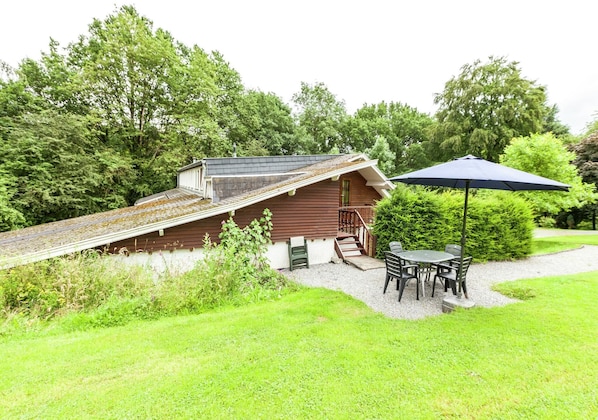 Eine Holzhütte mit geneigtem Dach, einer kleinen Veranda und einem Tisch mit Stühlen unter einem blauen Sonnenschirm.