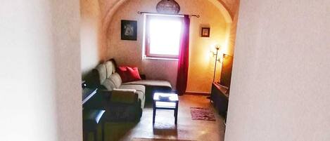 Un couloir étroit et voûté mène à un salon où se trouvent un canapé, une petite table et une lampe.