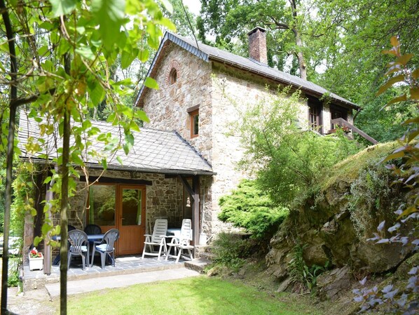 Ein Steinhaus mit einer überdachten Terrasse, Außenbestuhlung und einem üppigen Garten.