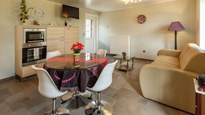 Un salon moderne comprenant un canapé, une table à manger avec deux chaises, un coin cuisine avec un four et un téléviseur, ainsi qu’un luminaire fixé au plafond.