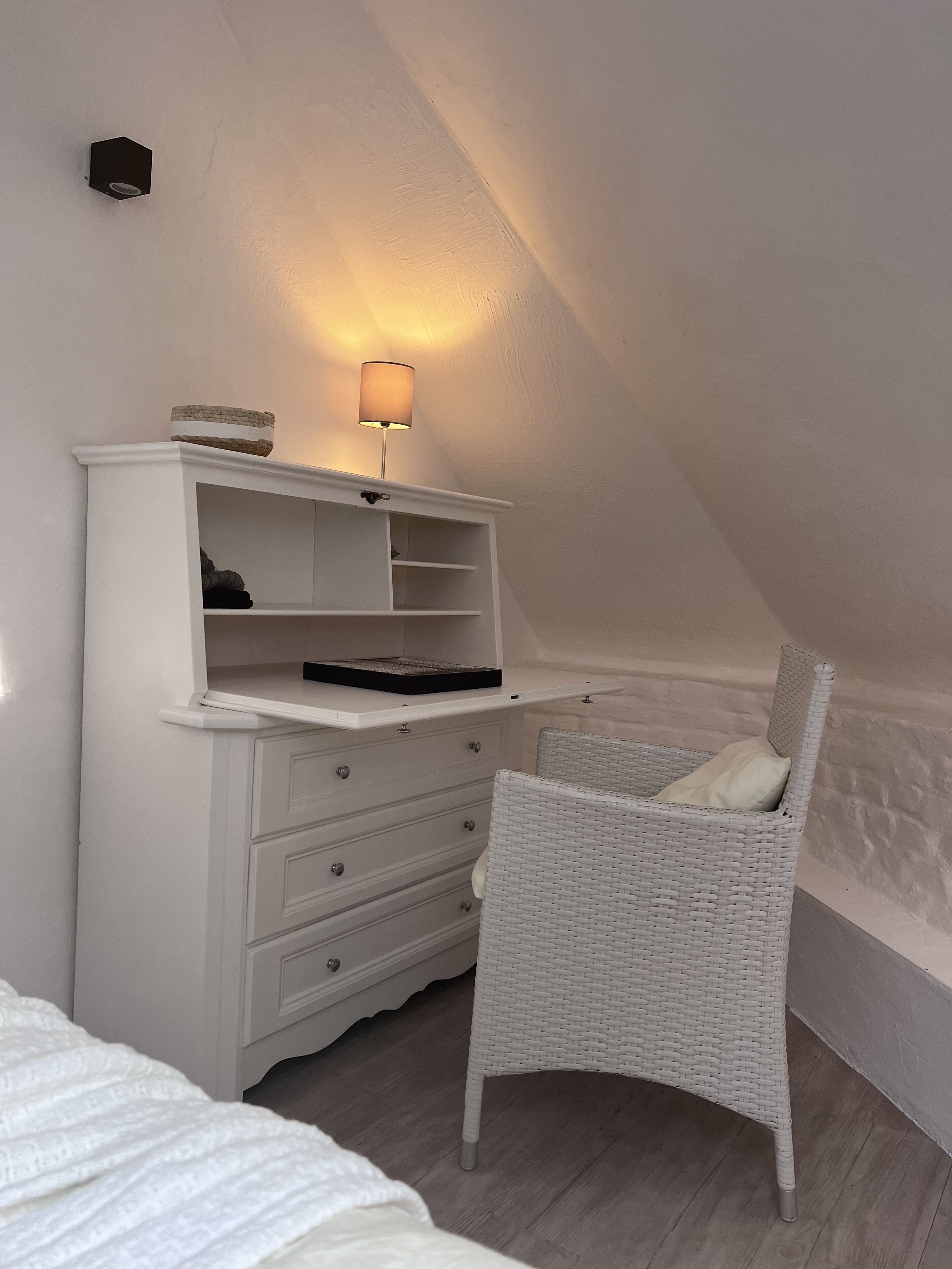 Um quarto estilo loft branco, com teto inclinado, cômoda branca com gavetas, cadeira de vime e mesa de cabeceira com abajur.