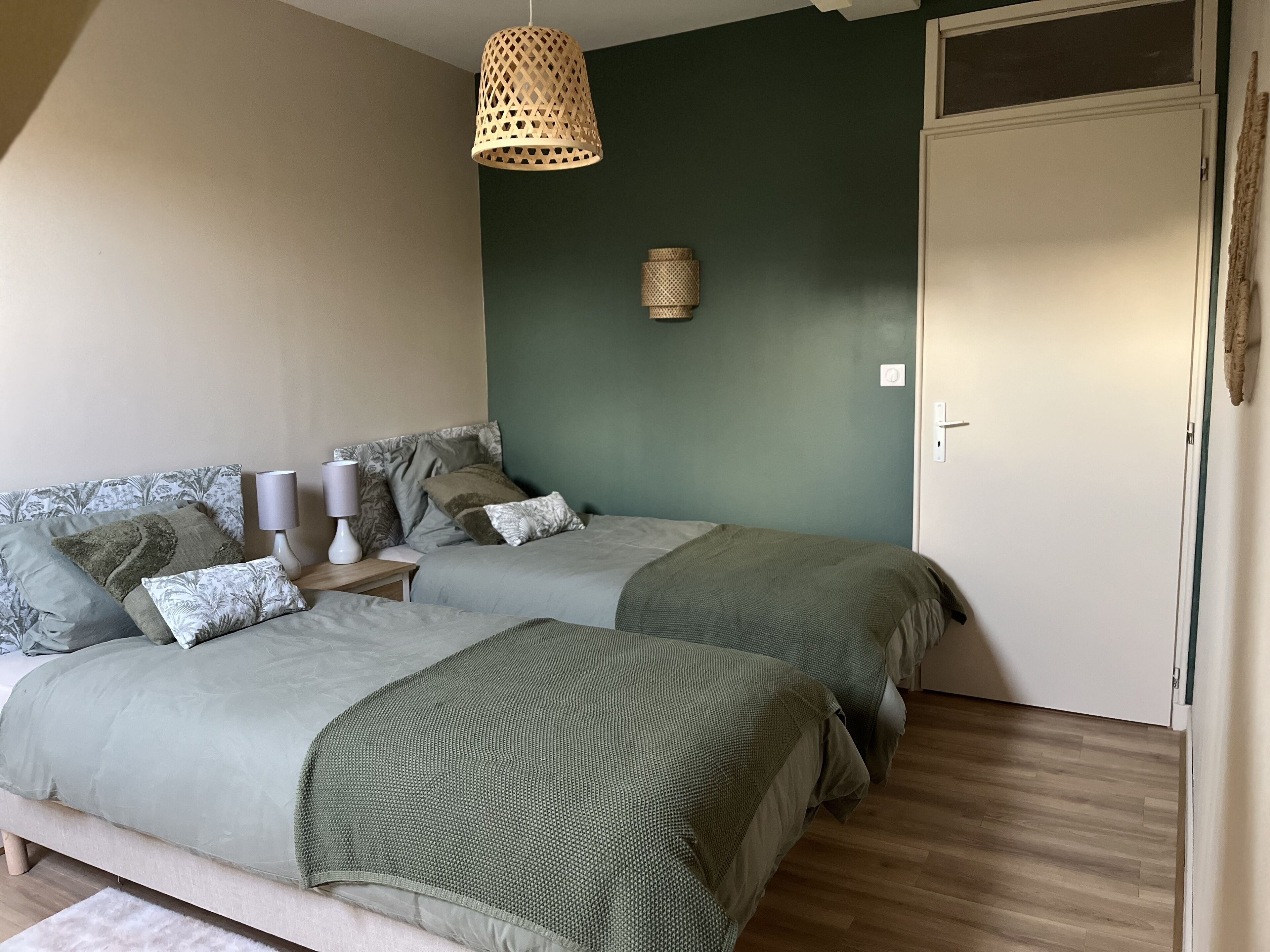 A bedroom with two beds, a hanging light fixture, a bedside table with a lamp, and a door.