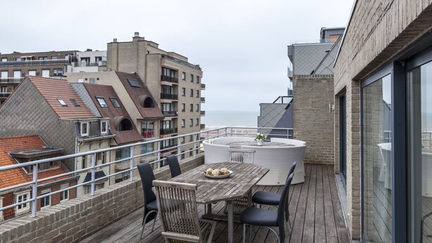 A rooftop terrace with a round table and chairs, overlooking a cityscape and the sea.