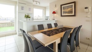 A modern kitchen with white cabinets, a dining area with a wooden table and black chairs, a red floor lamp, and a framed painting on the wall.