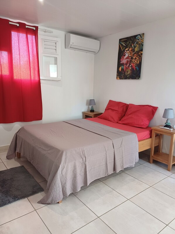 Une chambre à coucher avec un lit, une table de chevet, un climatiseur, une fenêtre avec un rideau et un tableau au mur.