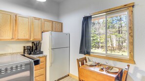 A compact kitchen with wooden cabinets, a stainless steel refrigerator, a coffee maker, and a dining table set for two with chairs. - Townsend Cottage on UTV & Snowmobile Trail! (Townsend)