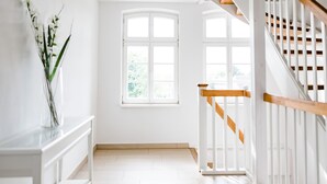 Una habitación luminosa y blanca con un ventanal grande, una escalera de madera y un banco blanco con un jarrón de flores.