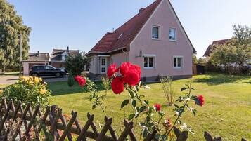 Une maison rose avec un toit rouge, entourée d’une clôture en bois et agrémentée de fleurs rouges.