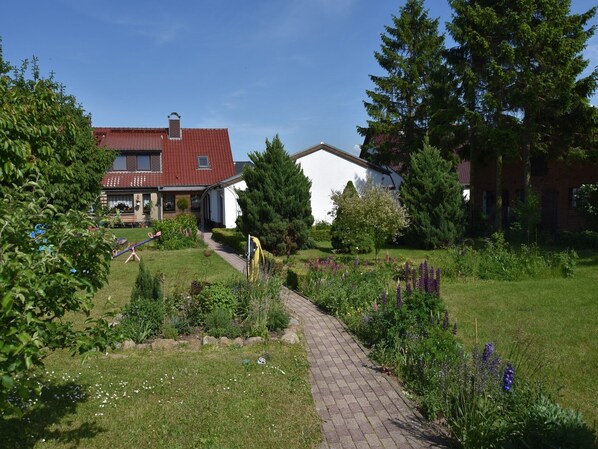 A garden pathway leading to a house with a red roof, surrounded by various plants and trees.