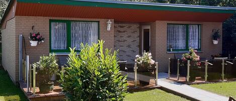 A small brick building with a green roof, two windows with white curtains, and a small porch area with potted plants.