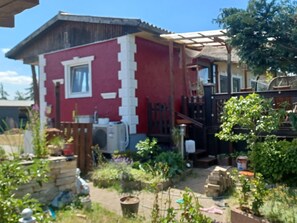 Un edificio rojo y blanco con una ventana y una puerta, rodeado de follaje y varios objetos.
