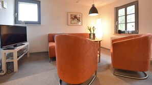 Een moderne woonkamer met een flatscreen televisie, oranje fauteuils, een kleine tafel met een lamp en een raam met uitzicht.