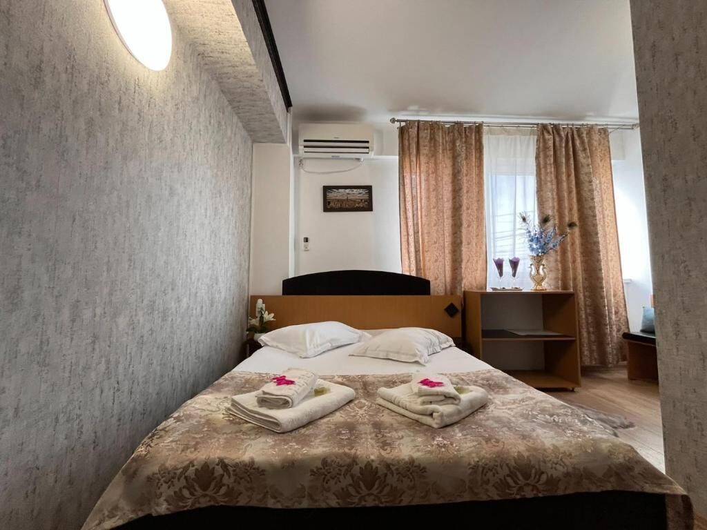 A hotel room with a bed, a nightstand, a window with curtains, and an air conditioning unit.