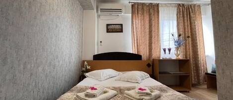 A hotel room with a bed, a nightstand, a window with curtains, and an air conditioning unit.