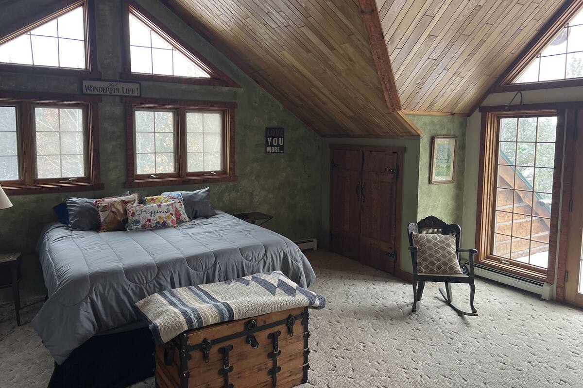 A bedroom with a lofted ceiling, large windows, a bed with multiple pillows, a chest of drawers, and a rocking chair.