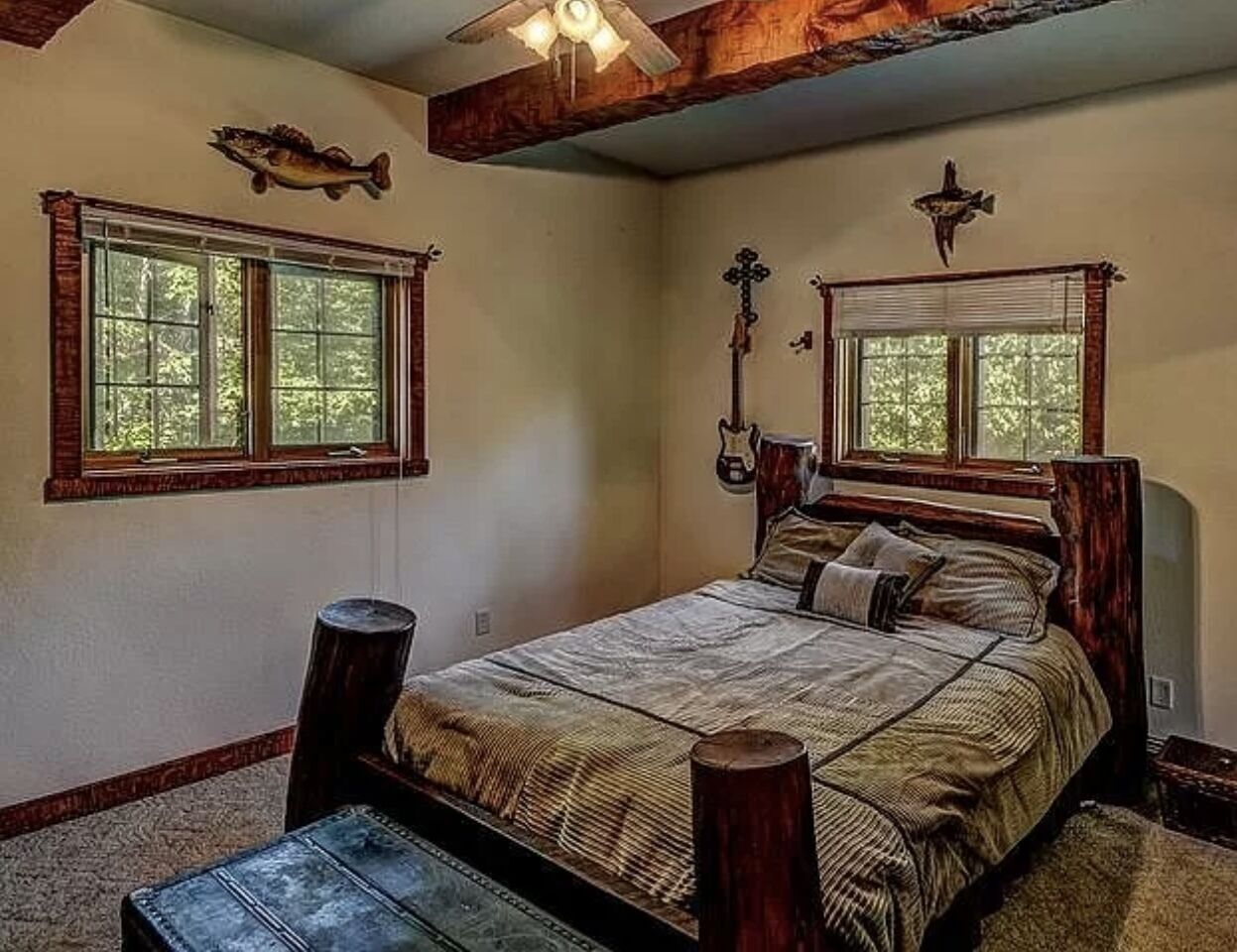 A bedroom with a large bed, two windows, wooden beams, and a ceiling fan.