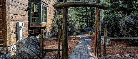 A wooden cabin with a stone foundation, a paved pathway leading to a wooden archway, and a forested backdrop.