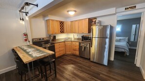 Cocina compacta con armarios de madera, un refrigerador de acero inoxidable, un microondas y una zona de comedor con mesa y taburetes de bar.