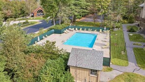 Un área de piscina con sillones de descanso y un pequeño edificio, rodeado de árboles y vistas a la montaña.