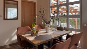 Ein Essbereich mit einem Holztisch, Stühlen, einer Vase mit Zweigen und einem Fenster mit Blick auf ein Holzgebäude.