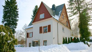 Uma casa de dois andares com telhado vermelho e paredes brancas, rodeada por neve e árvores.