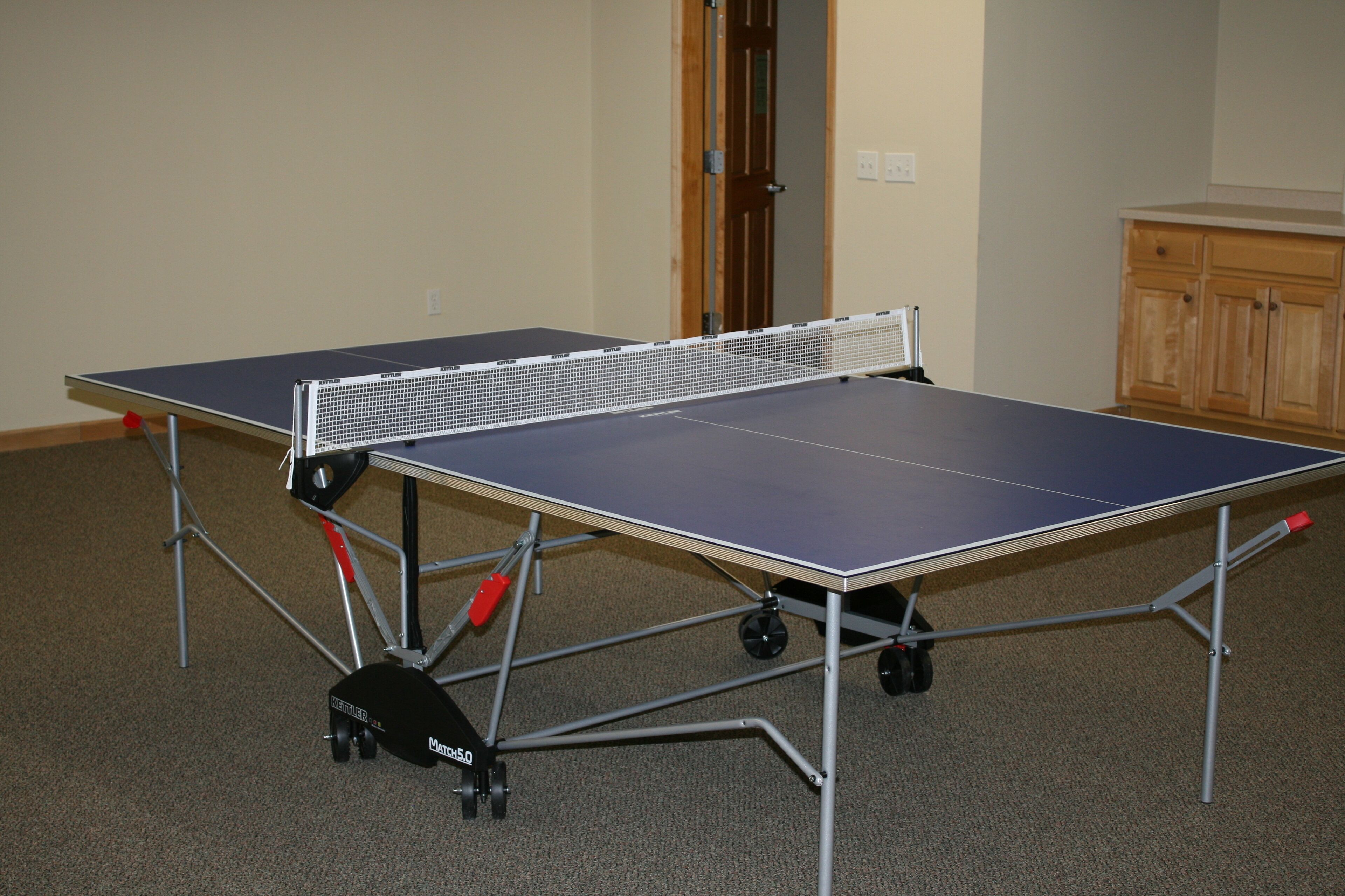 A table tennis table with wheels, a door, and a wooden cabinet in the background.