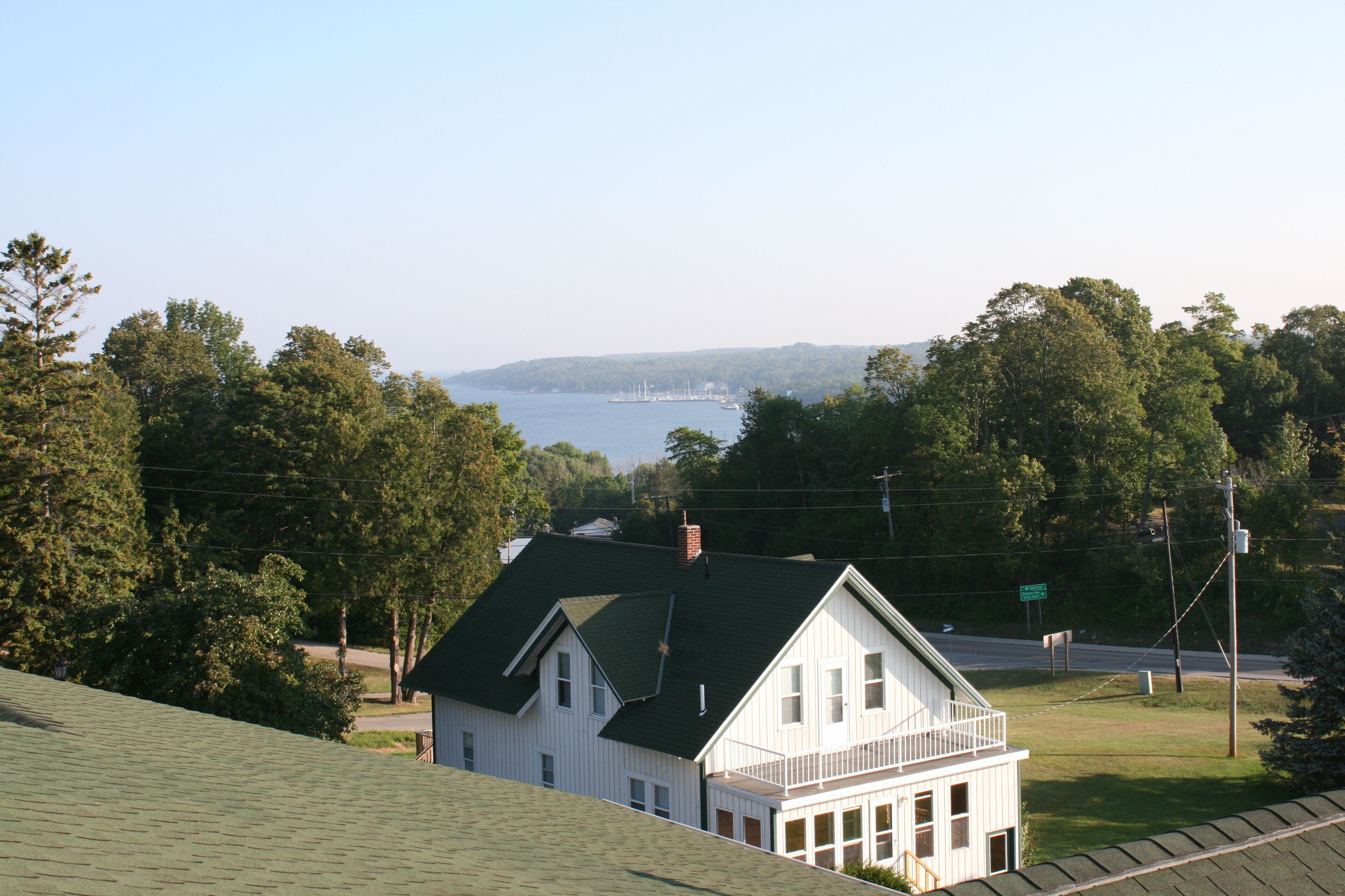 A white house with a green roof, a balcony, and a view of a body of water and trees in the background.