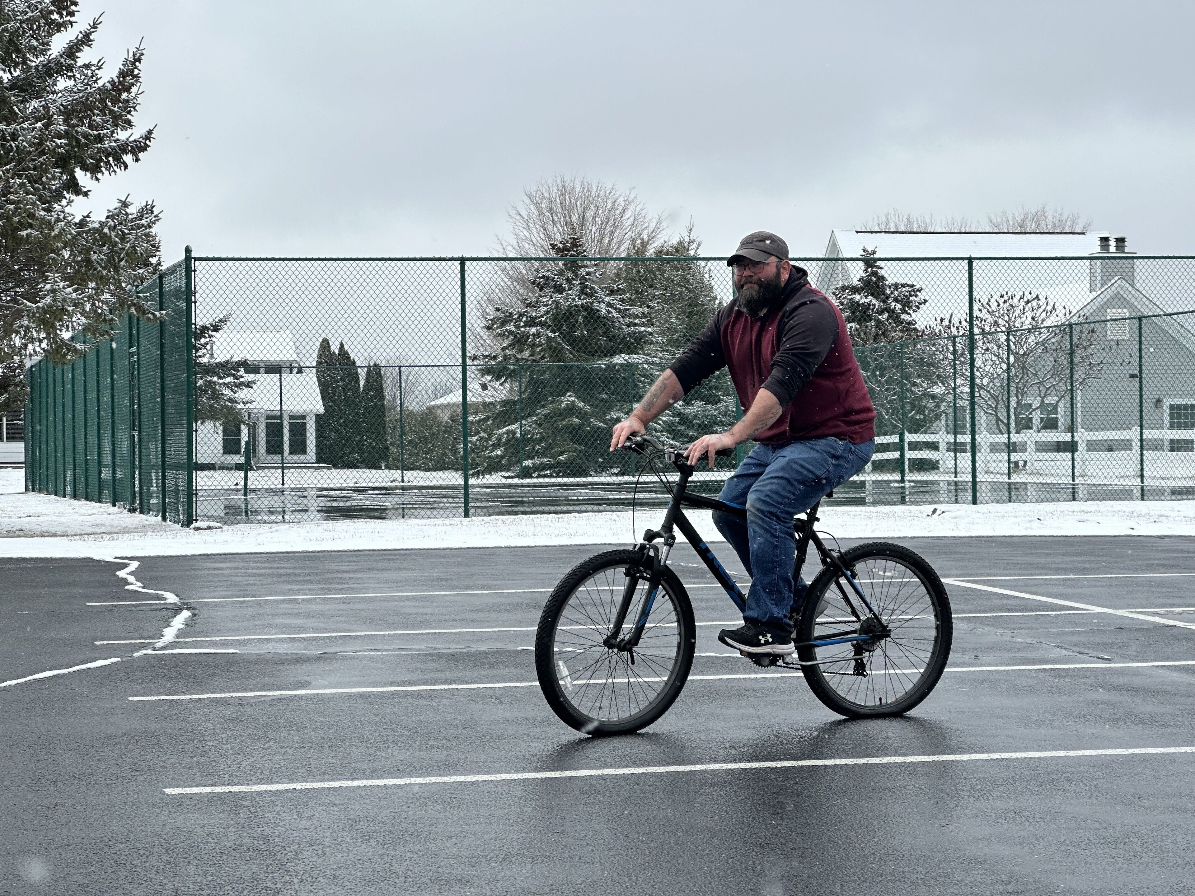 A person riding a bicycle on a paved surface with a fence and trees in the background.