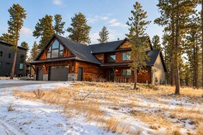 A two-story log cabin with a garage, surrounded by tall pine trees and a snowy landscape.