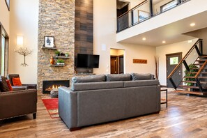 A spacious living room with a stone fireplace, a grey sectional sofa, a flat-screen TV, and a staircase with a glass railing.