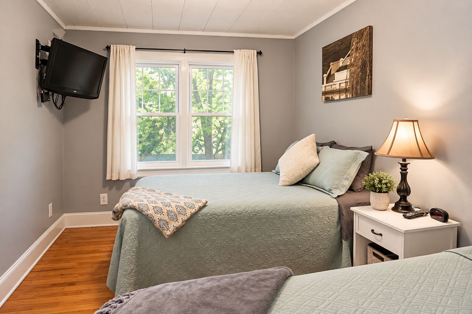 A bedroom with two beds, a TV mounted on the wall, a window with curtains, a bedside lamp, and a painting on the wall.
