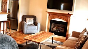 Una sala de estar con chimenea, televisor y una zona de cocina con refrigerador.