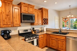 A kitchen with wooden cabinets, a stainless steel refrigerator, a microwave, a stove, a dishwasher, and a window with a view of greenery.