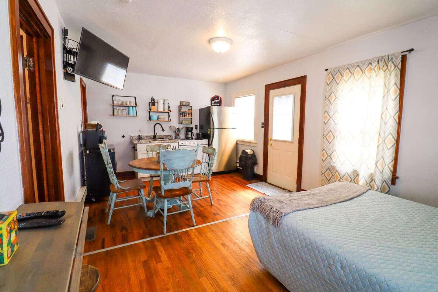 A compact room with a bed, a small kitchenette, a dining table with chairs, and a TV mounted on the wall.