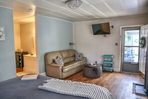 A living room with a beige sofa, a grey ottoman, a flat-screen TV, and a door leading to an outdoor area.