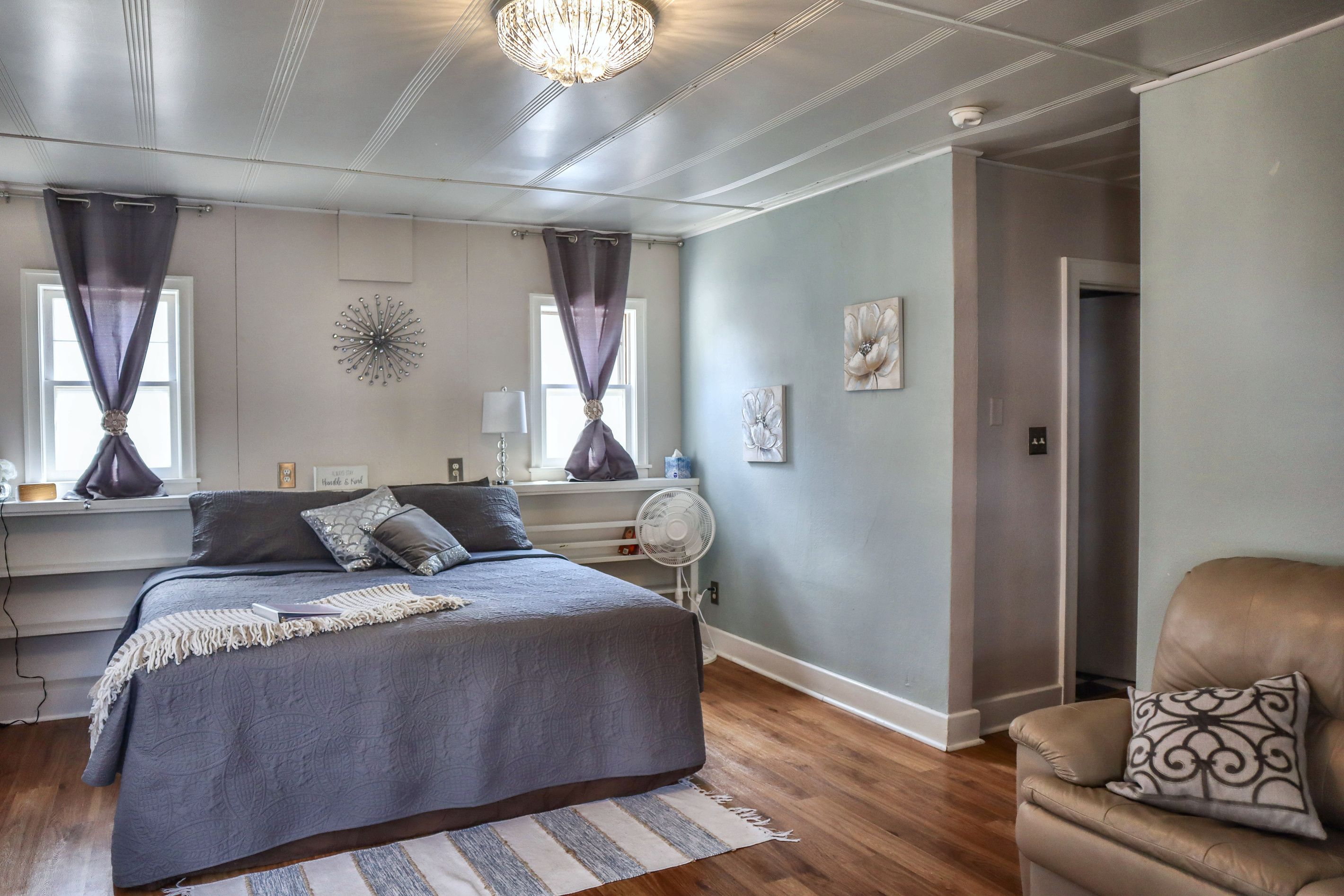 A bedroom with a bed, a chair, a fan, and a window with curtains.