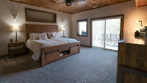 Une chambre avec un plafond en bois, un lit doté de tiroirs de rangement, une table de chevet, une lampe et une fenêtre avec des stores.